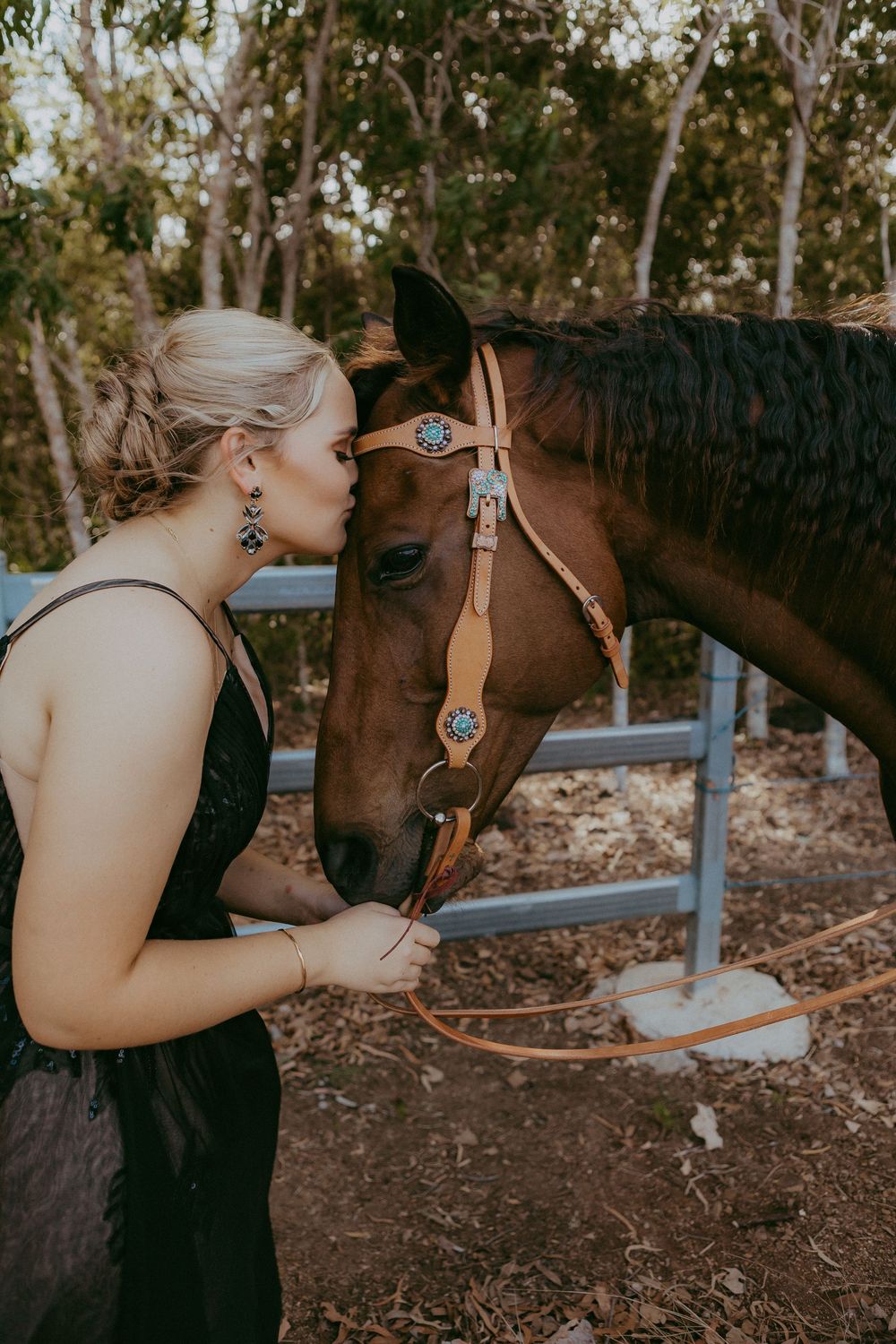 Blonde woman in black dress kissing chestnut horse wearing turquoise bridle in wooded area.
