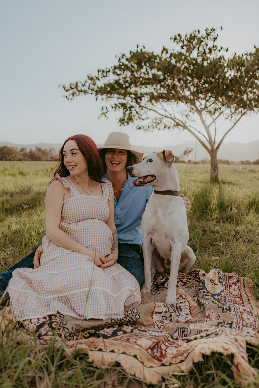 Pregnant woman and partner laugh together with white dog on blanket in sunny green field.