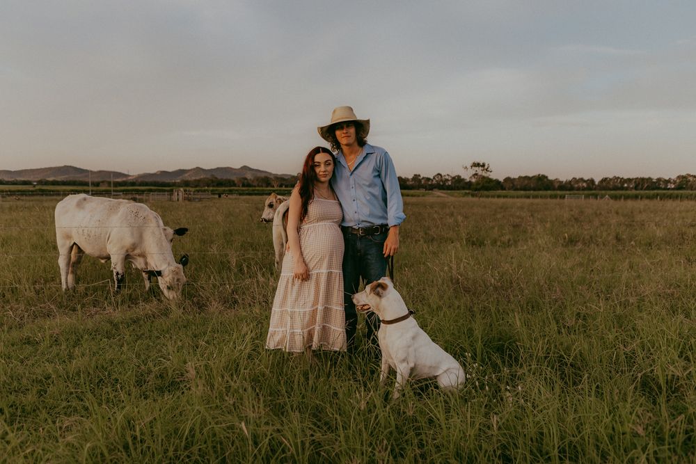 Couple with dog and cow in green pasture at golden hour, farm lifestyle maternity photo session.