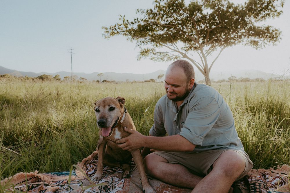 Man in casual shirt bonds with senior brown dog on patterned rug in sunlit grassy field.
