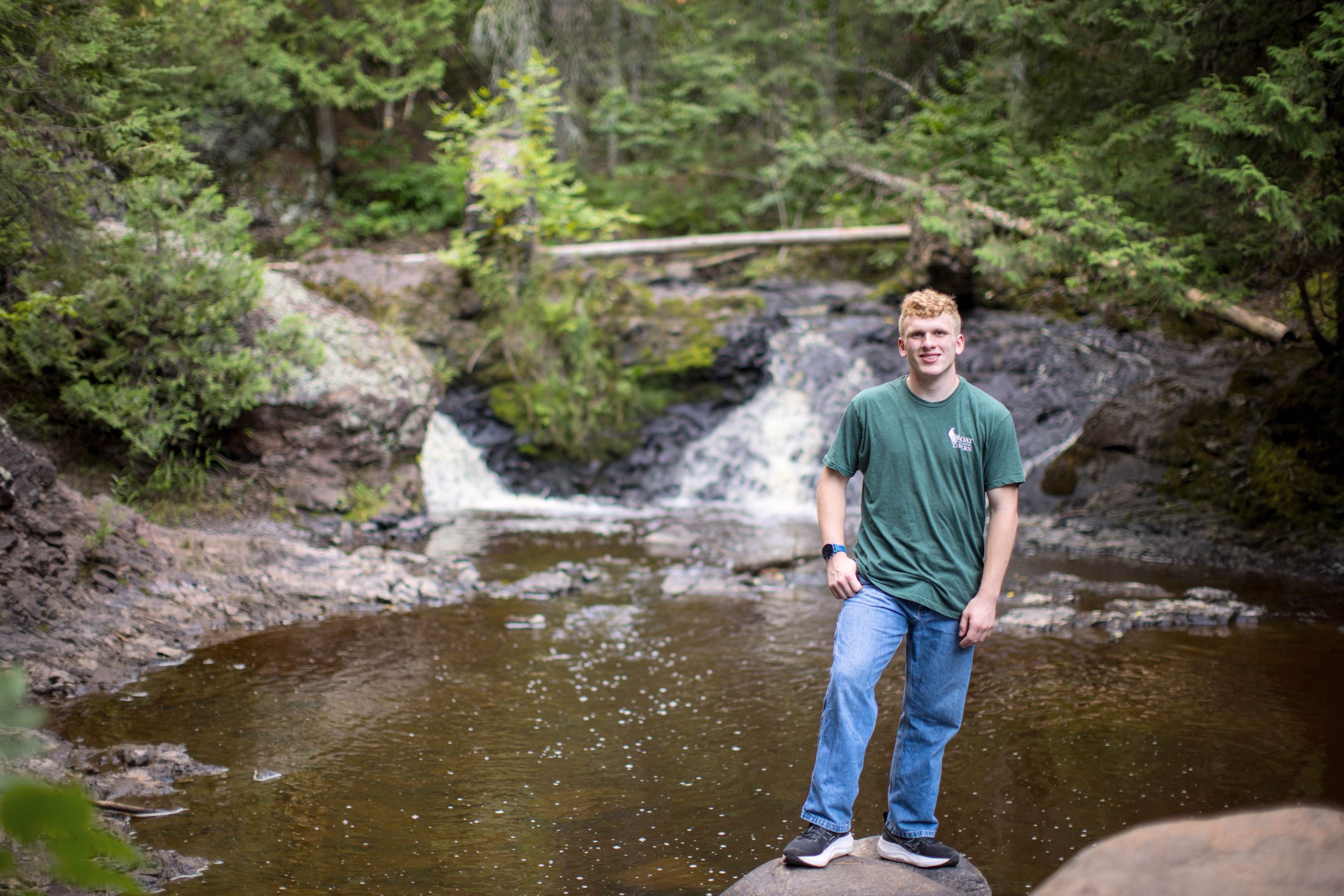 A scenic waterfall cascades behind someone standing on a rock in a forest stream.