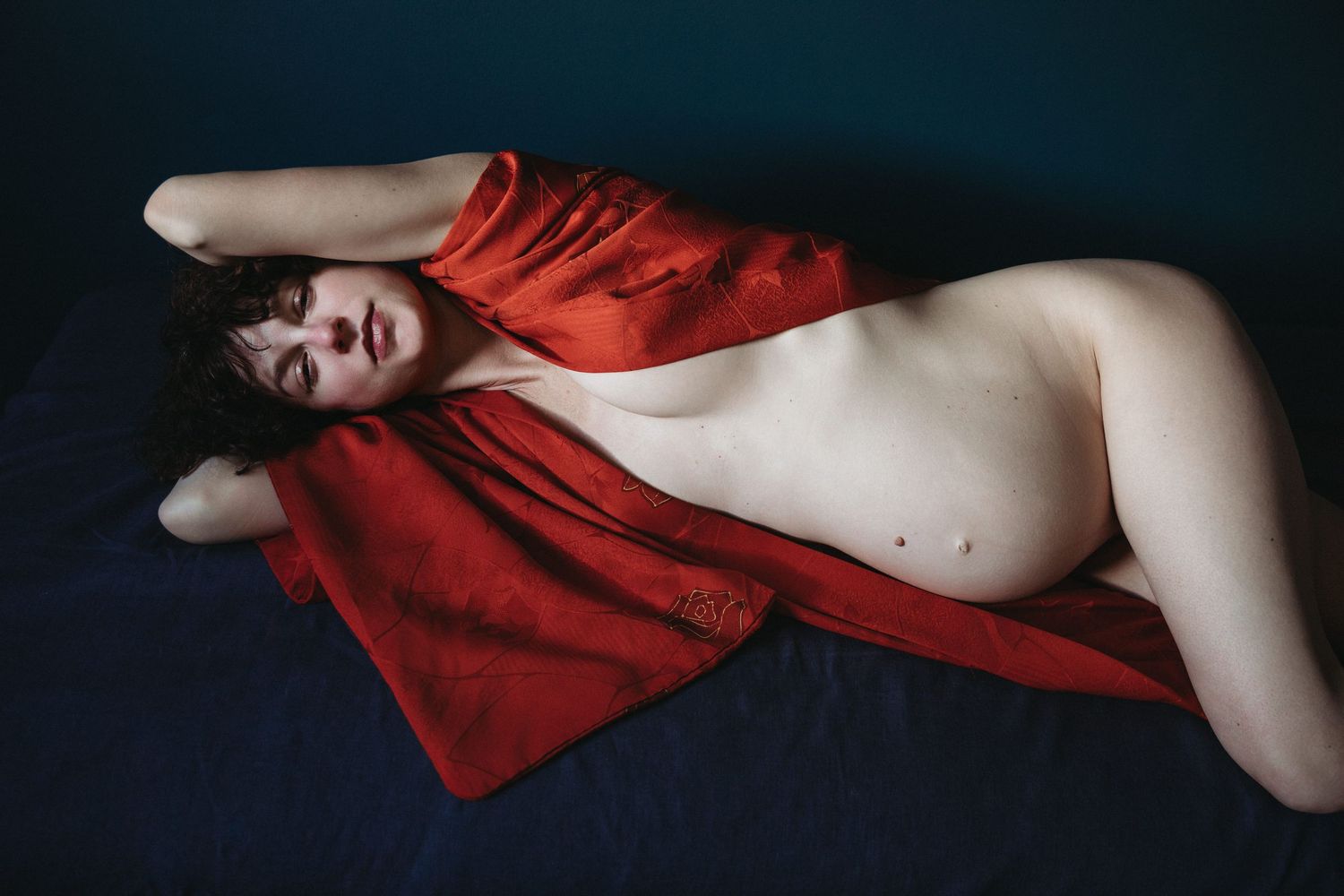 woman lies on back wearing red robe and displaying bump for pregnancy photographer london sarah stirk