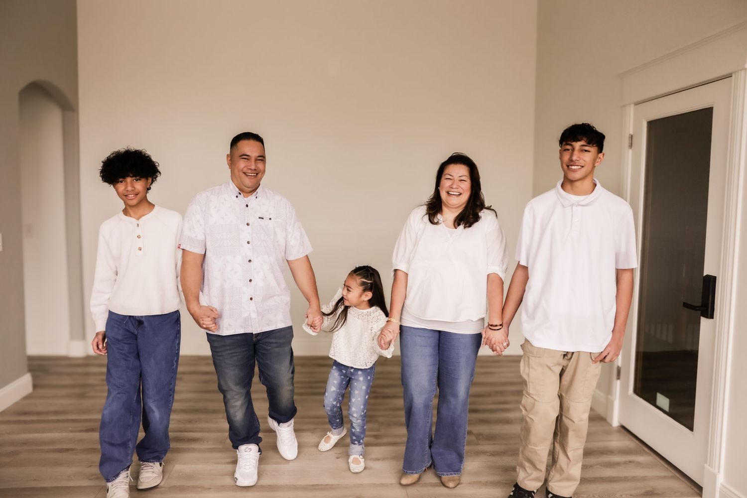 Happy family of five holding hands in white shirts and jeans posing in a bright modern home entryway.