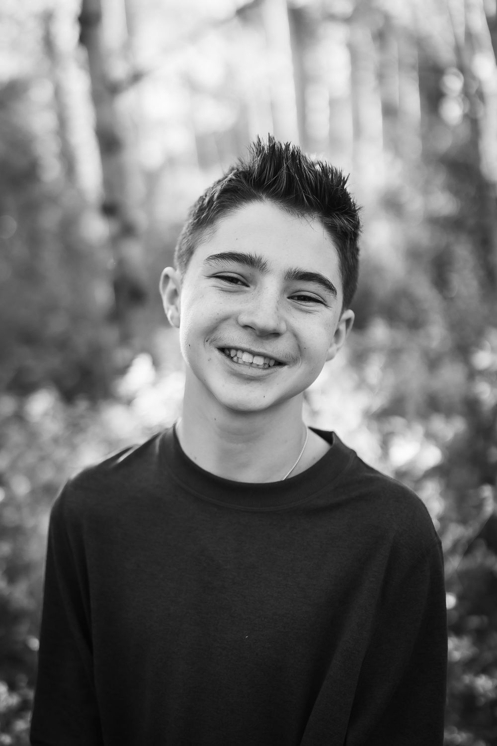 A black and white photo a child smiles broadly in front of trees in Colorado