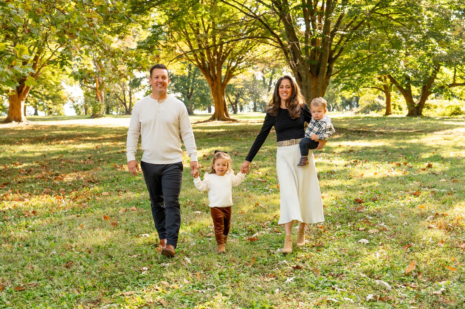 People holding their toddler outdoors on autumn day surrounded by fall foliage in park setting during seasonal photoshoot.