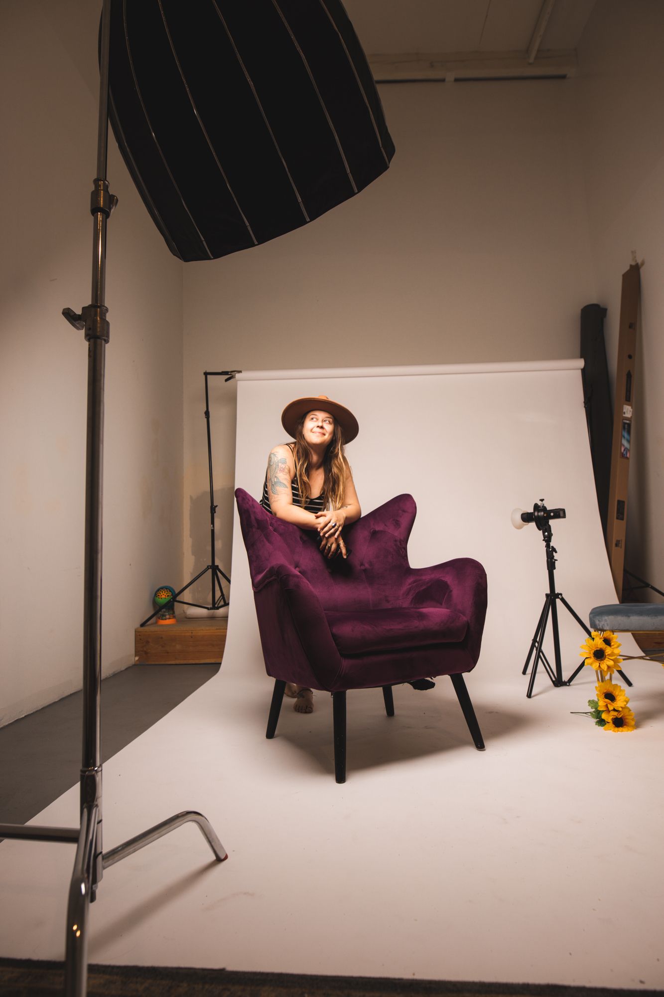 Female model in brown hat sitting on a purple velvet chair in a professional photography studio.