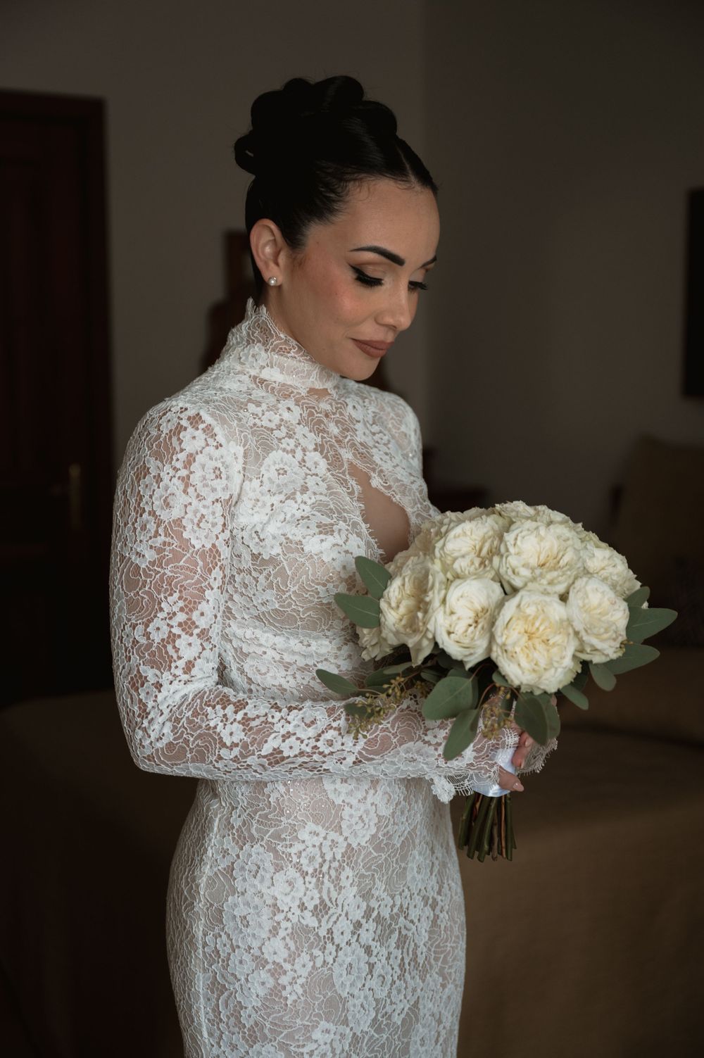Elegant bride in long-sleeve lace gown holding a bouquet of white roses against dark background.