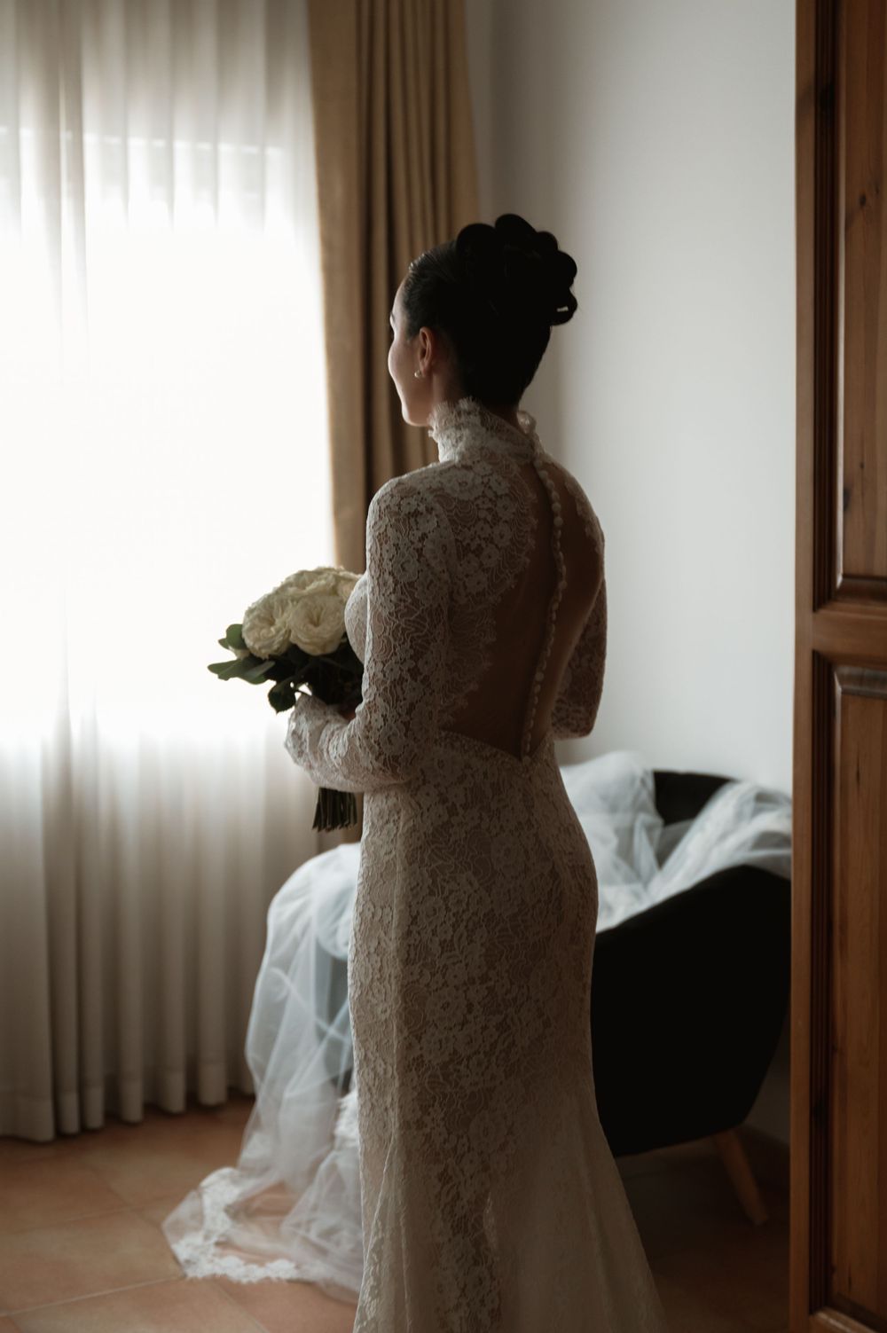 Bride in full lace dress stands by window holding white bouquet in natural light.