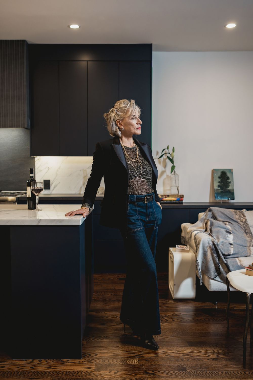 Elegant woman in black blazer and jeans leaning on kitchen island in modern dark-cabinetry home interior.