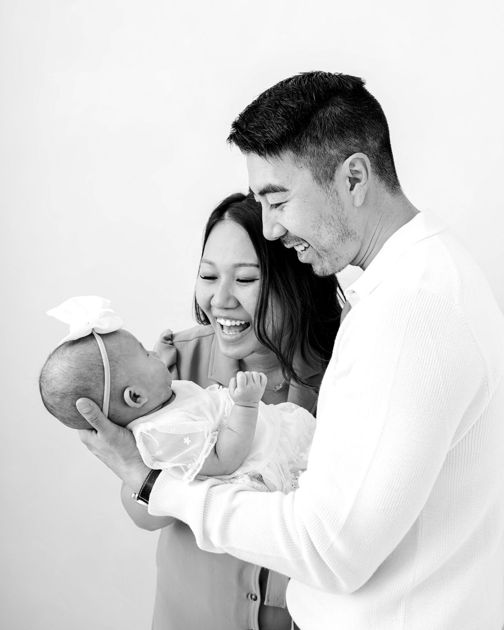 Black and white photo of smiling Asian parents lovingly holding their newborn baby with a white bow headband.