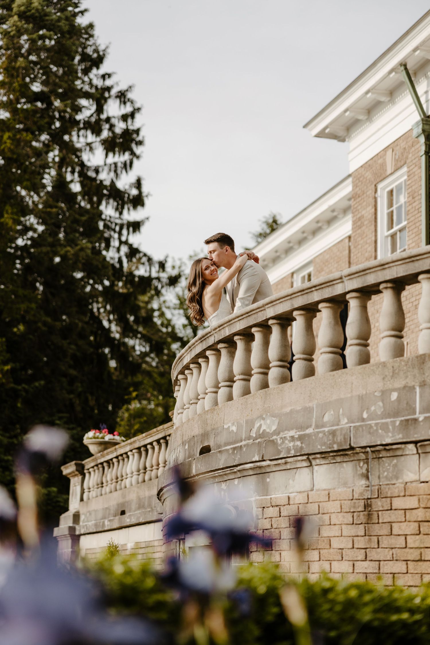 Couple embracing on a stone balcony of an elegant brick mansion surrounded by lush green trees. DFW engagement photographer