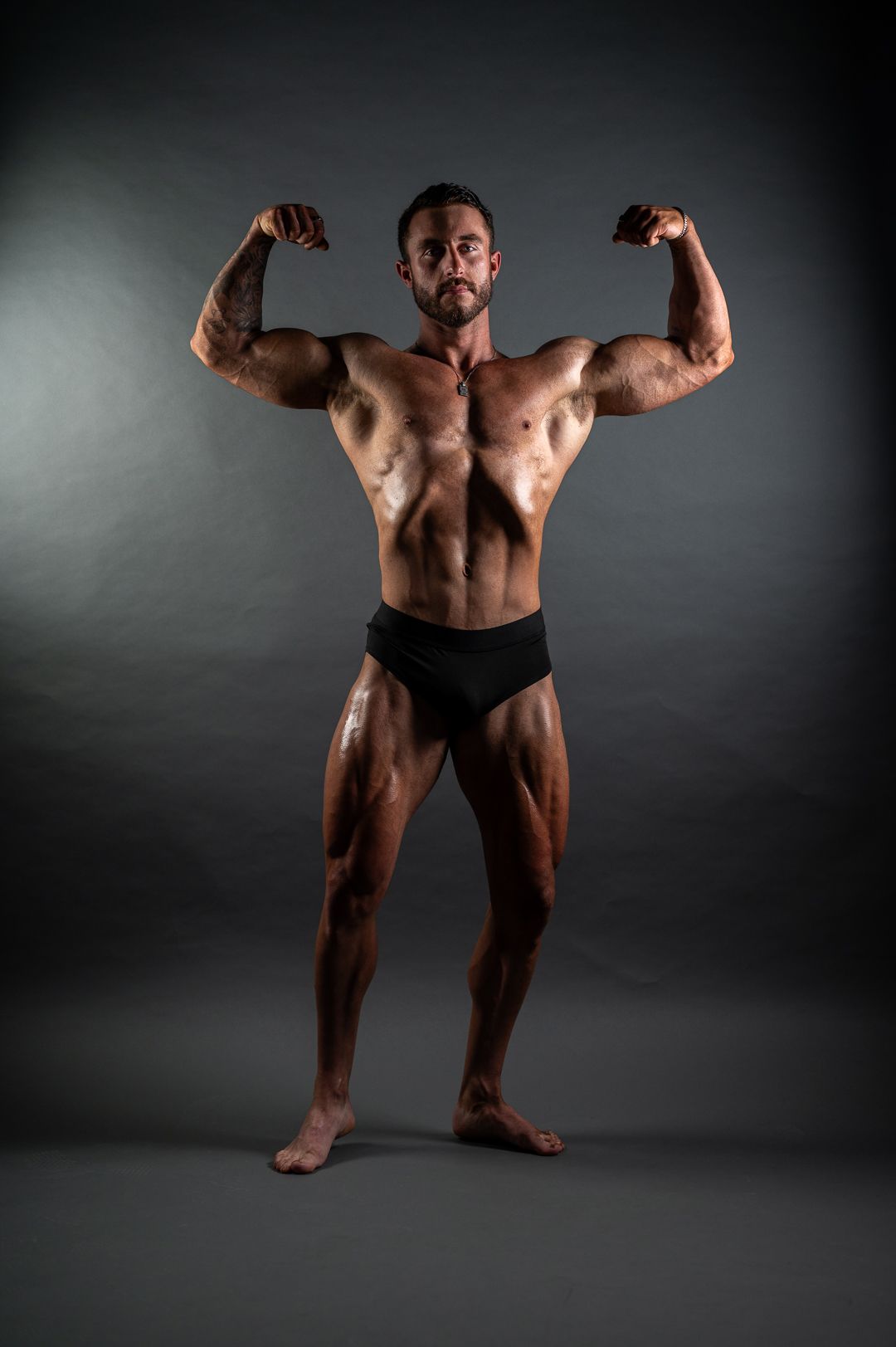 Muscular bodybuilder flexing biceps in a double front pose, wearing black briefs against a dark studio background.