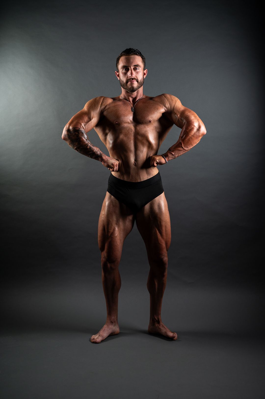 Muscular male bodybuilder posing in black briefs against gray background, showcasing defined muscles in classic competition stance.