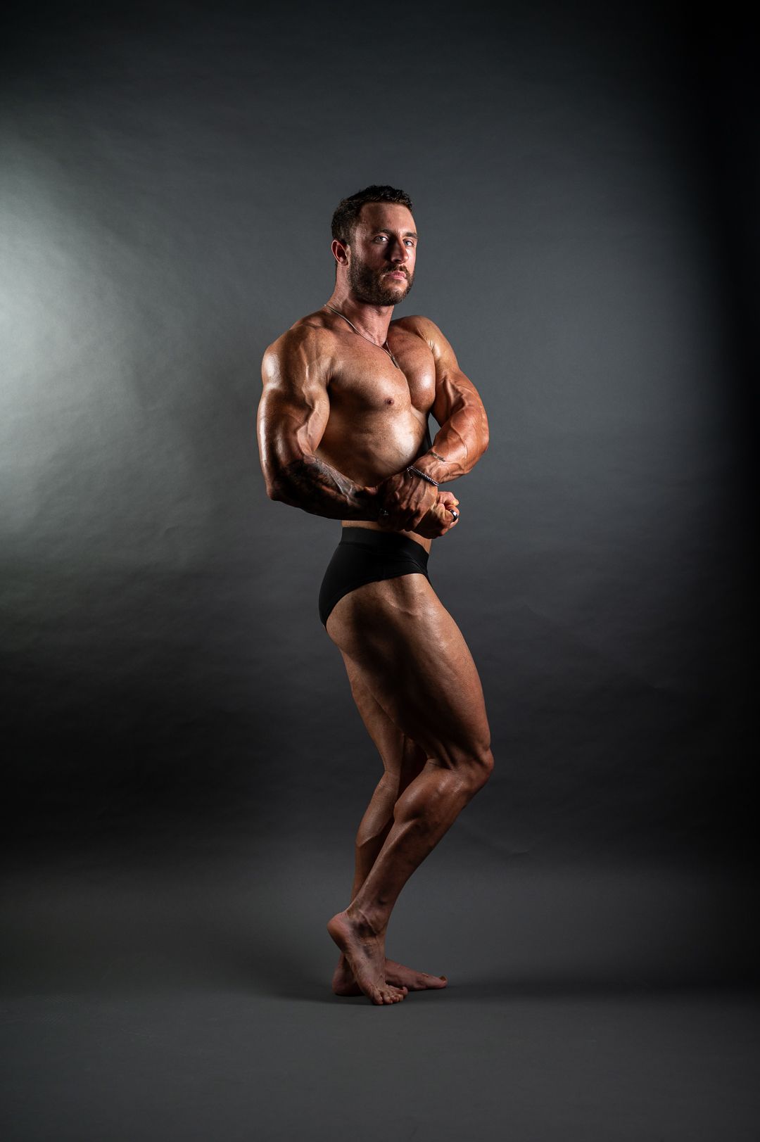 Muscular male bodybuilder posing shirtless in black shorts against dark studio background, showcasing defined physique.