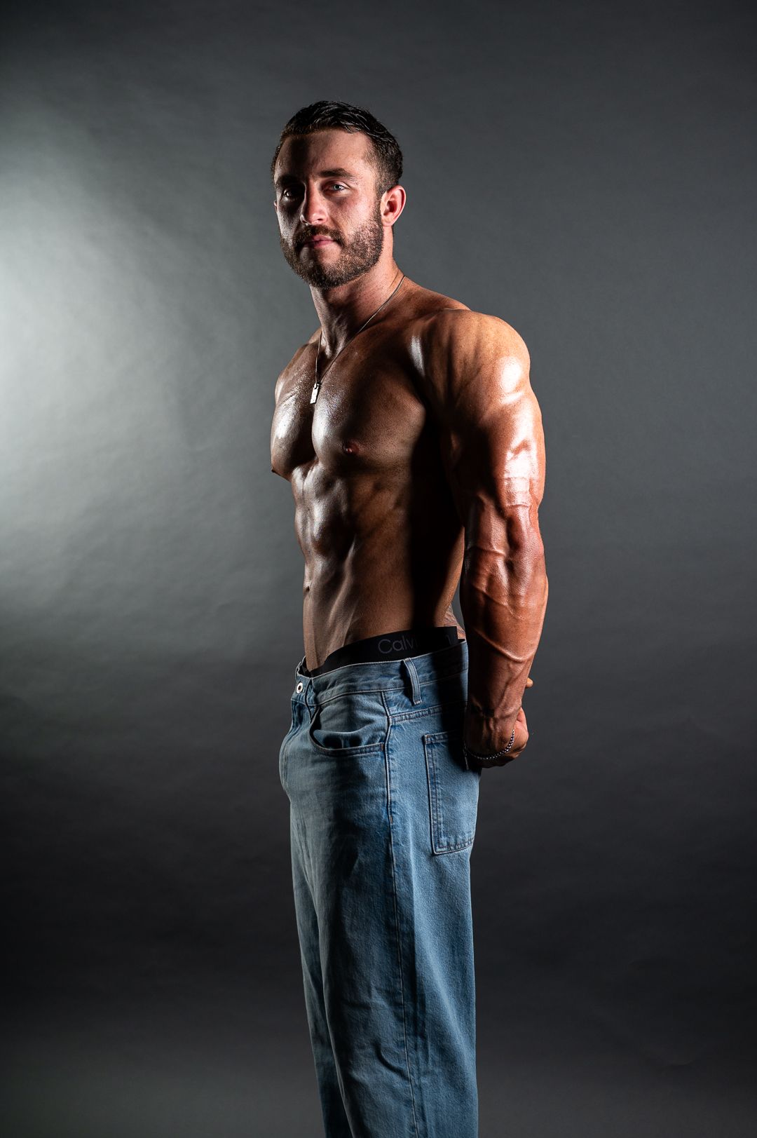Shirtless muscular man in jeans posing sideways showing defined abs and arms against dark background.