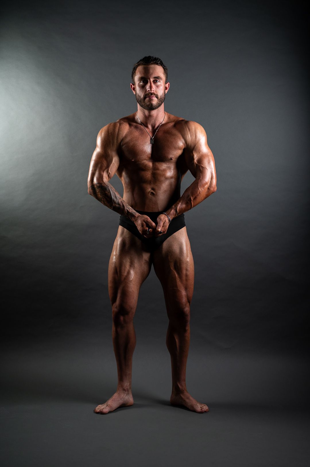Muscular male bodybuilder posing in black briefs against dark gray background showcasing defined physique.