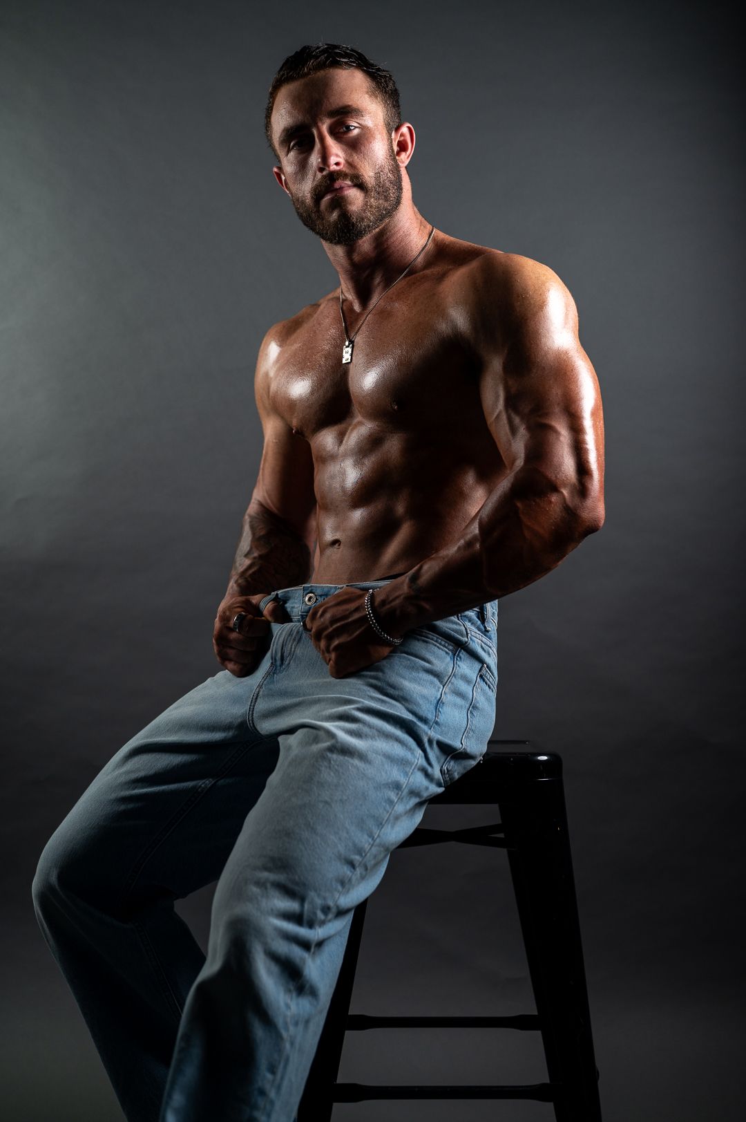 Muscular shirtless man sitting on a stool, wearing light blue jeans, showcasing defined abs and biceps against a dark background.