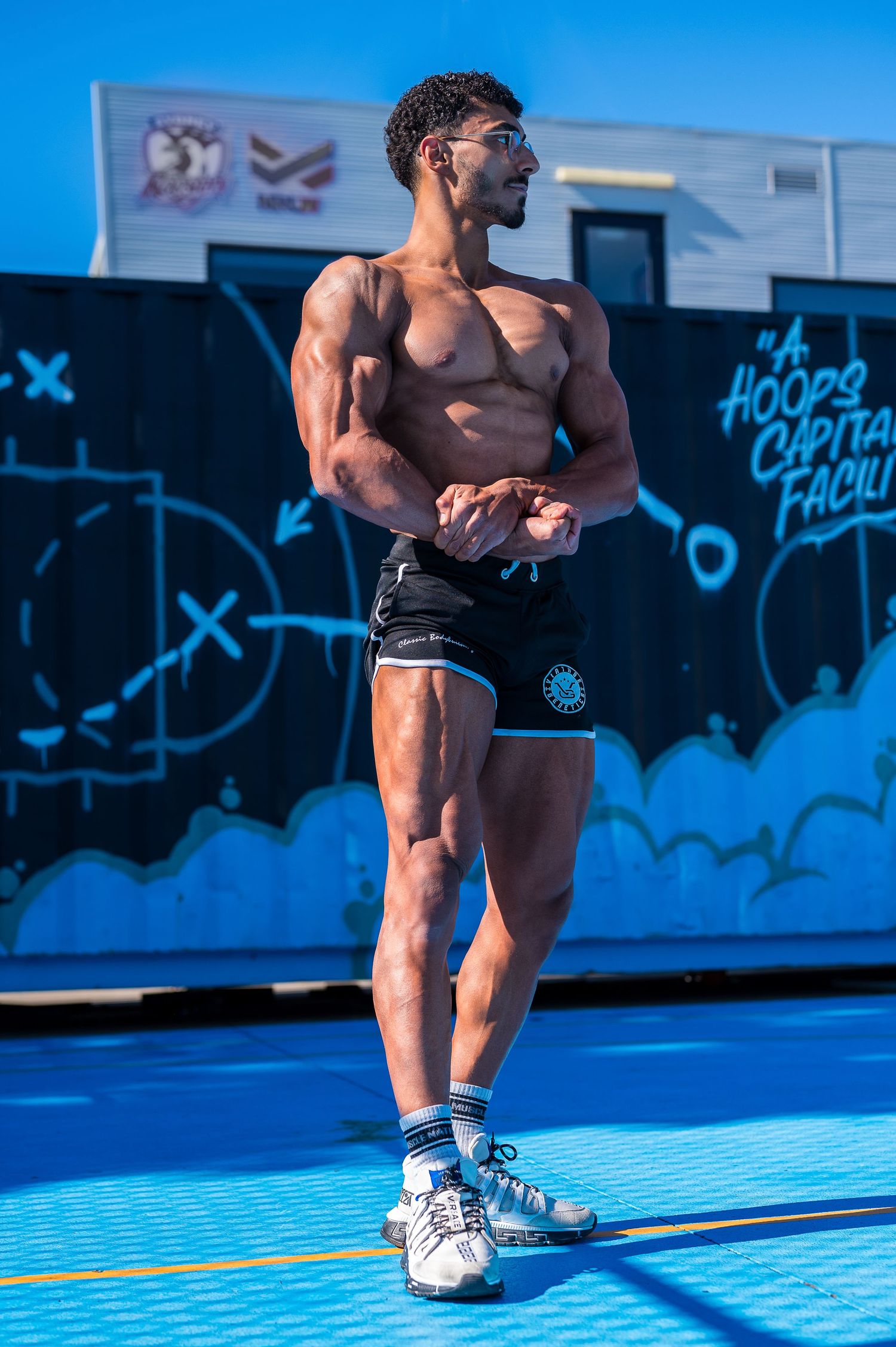 Muscular athlete posing outdoors against blue graffiti wall in athletic shorts and sneakers.