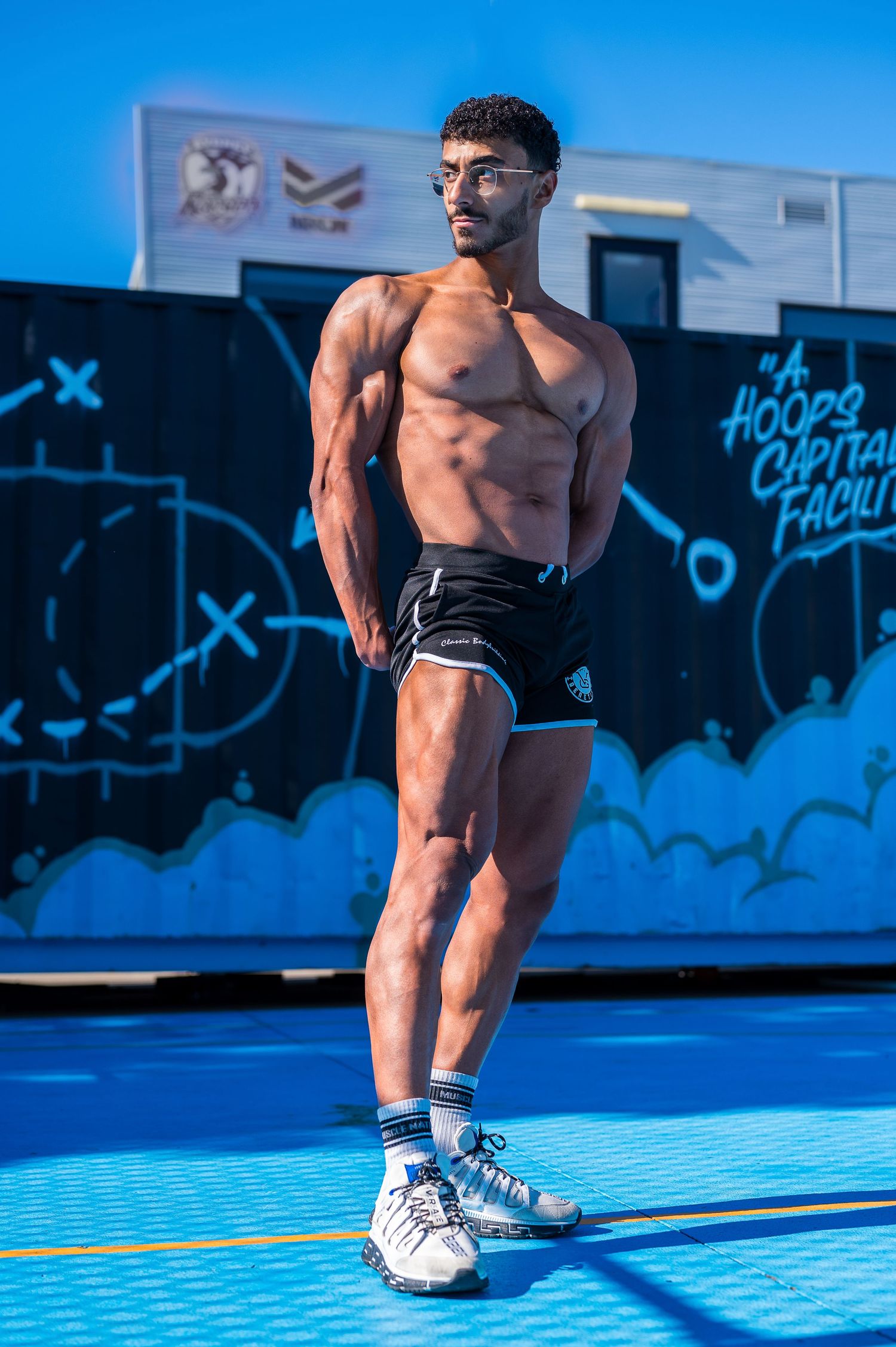 Fitness model in black shorts striking a pose against vibrant blue outdoor backdrop.