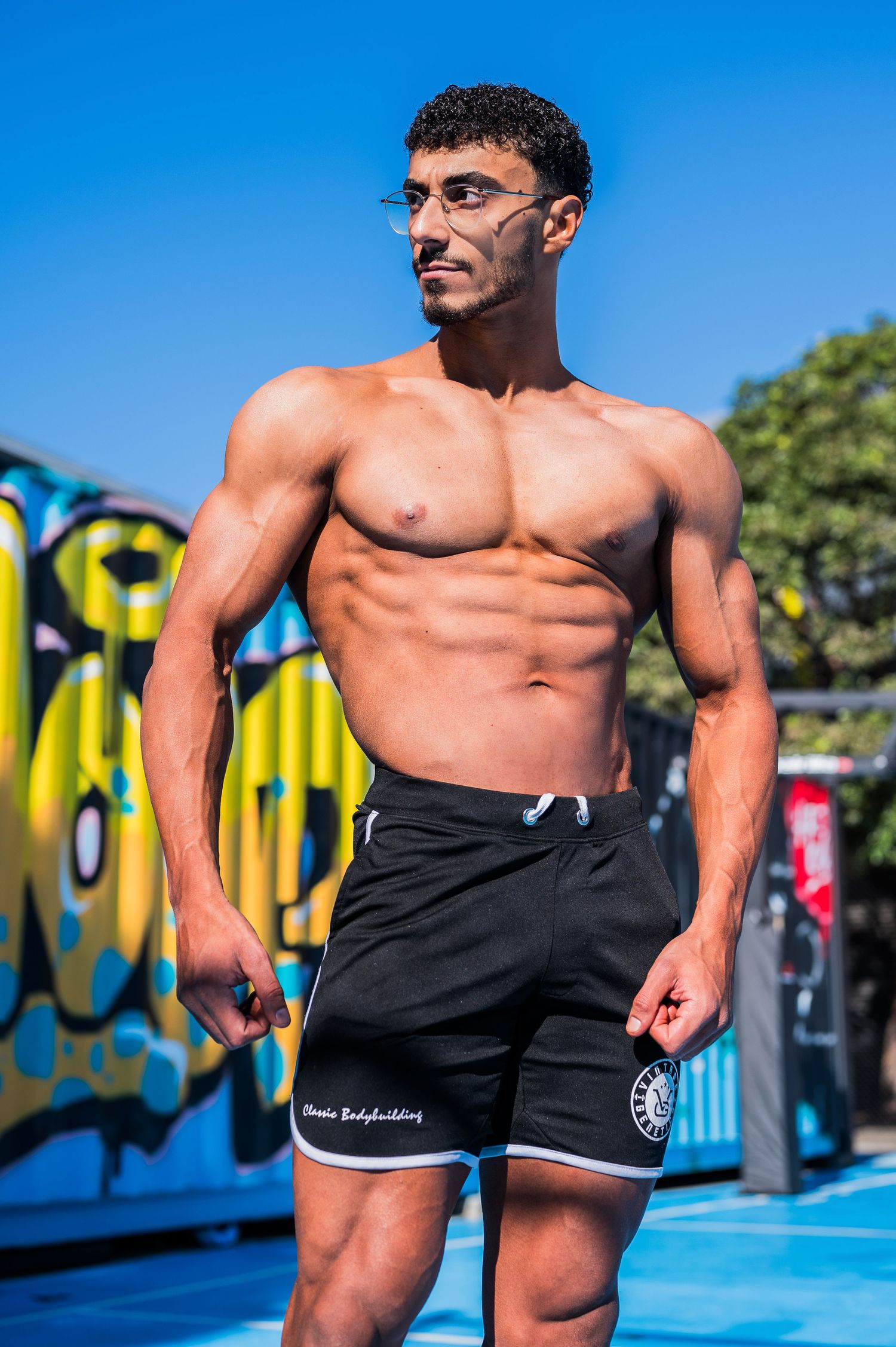 Muscular model in black athletic shorts posing outdoors with graffiti art backdrop.