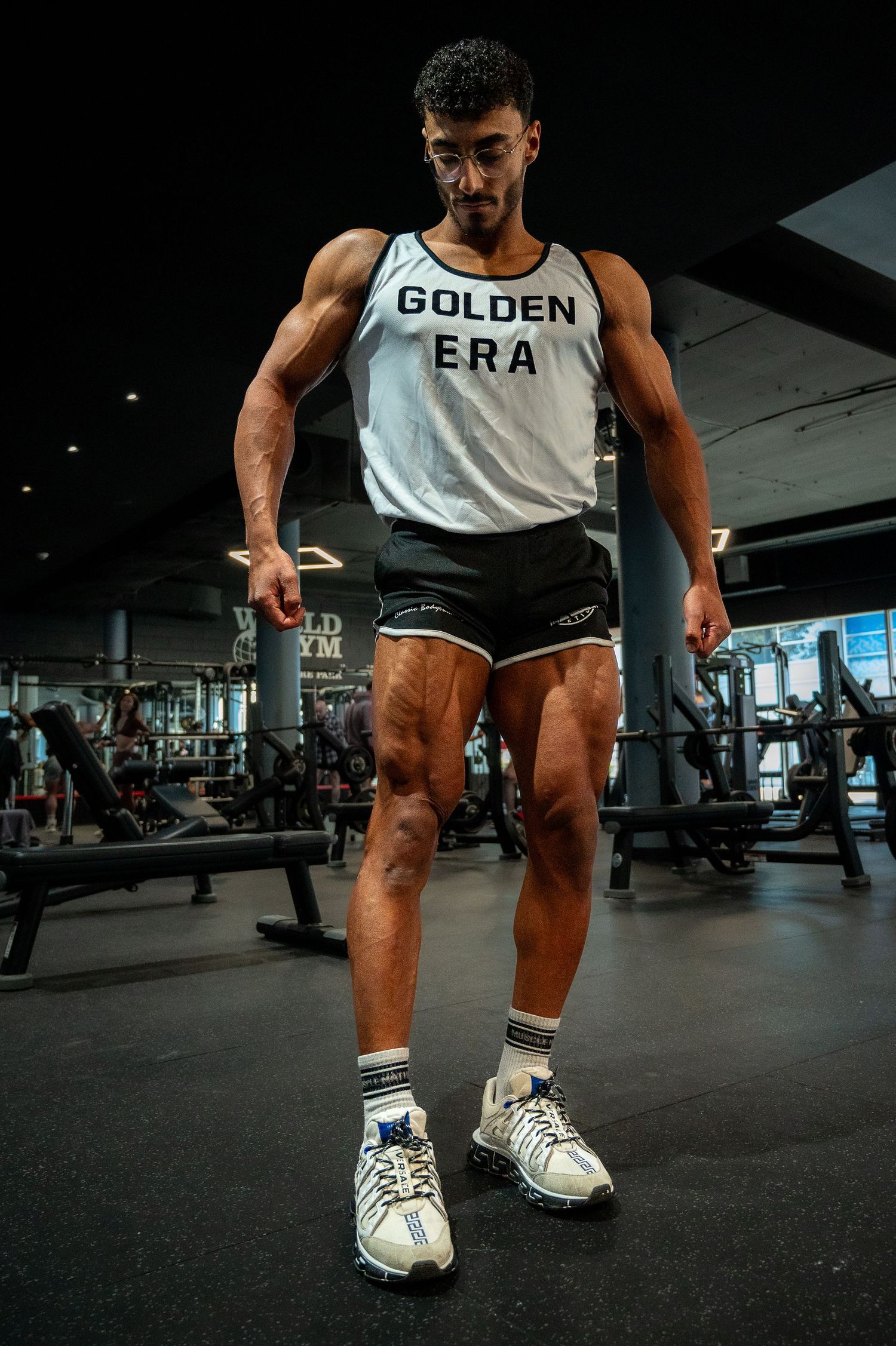 Fitness enthusiast in white tank top and shorts trains with weights in modern gym setting.