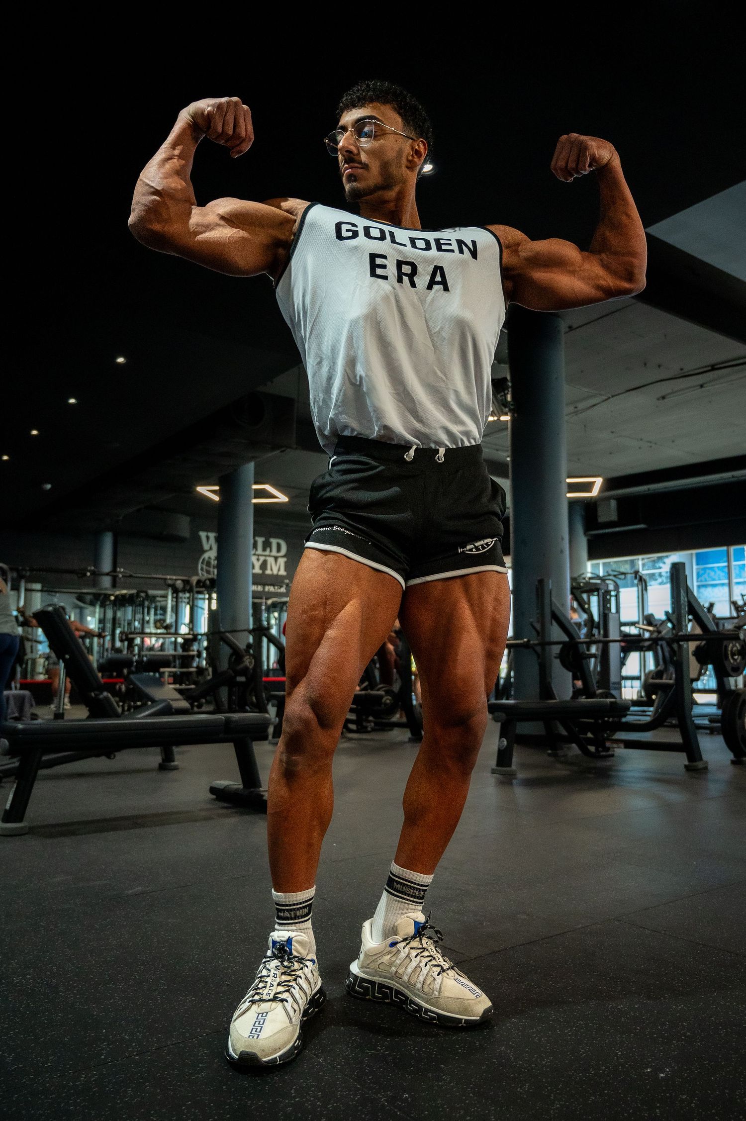 Athletic bodybuilder flexing muscles in a gym while wearing a 'Golden Era' tank top and shorts.