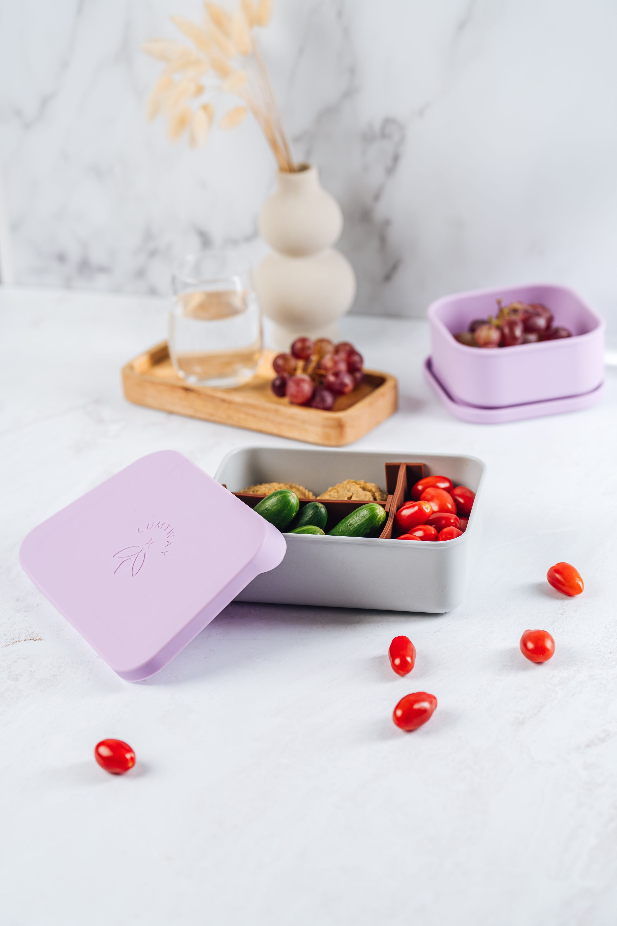 Healthy snacks and vegetables arranged in white and purple containers with cherry tomatoes scattered on marble surface.