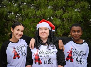 Three people wearing matching Christmas tribe shirts smile together against an evergreen background.