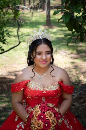 A person wearing a red quinceañera dress with gold details and a tiara poses in a wooded outdoor setting.