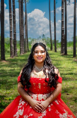 A person in a red and white formal gown sits on grass in front of tall pine trees on a sunny day.