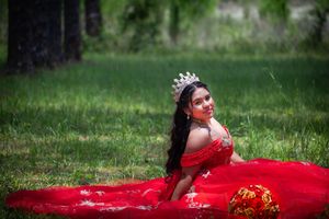 A person in a flowing red ballgown dress poses gracefully on lush green grass during a photo session.