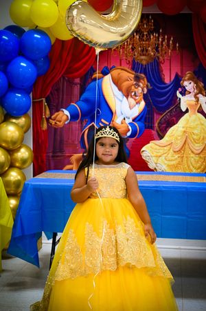 Young party guest in yellow princess dress poses with balloons and decorations at beauty and beast themed party event.