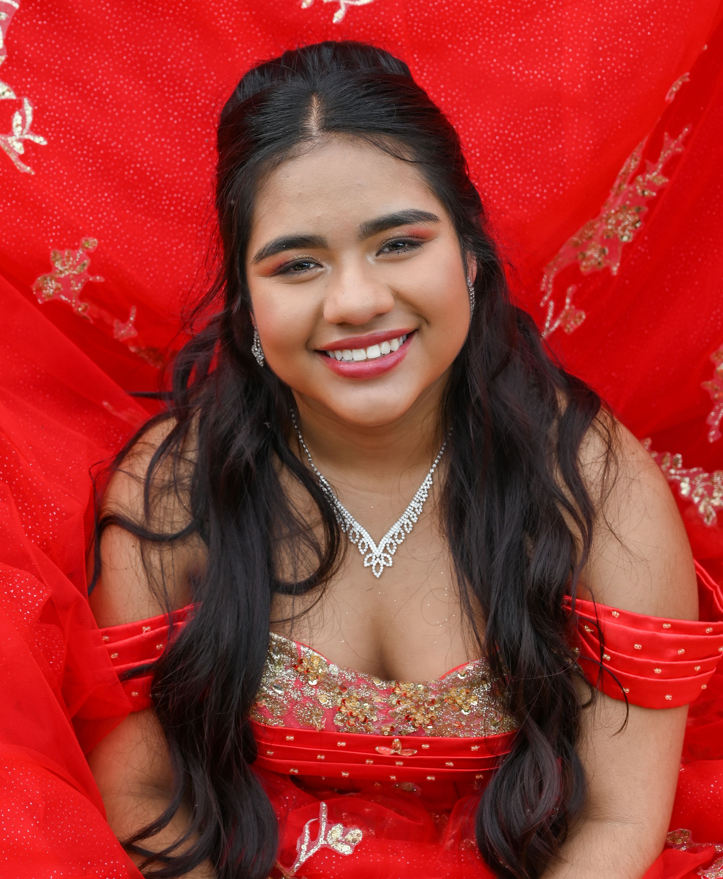 Young woman posing against vibrant red backdrop wearing traditional South Asian attire with ornate jewelry.