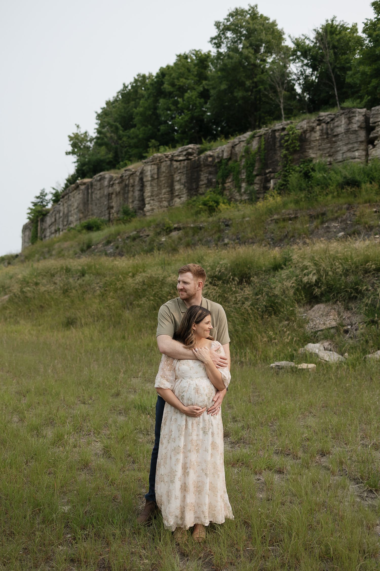 wife wrapped up in haubands arms infront of rock wall with green trees on top overland park photographer