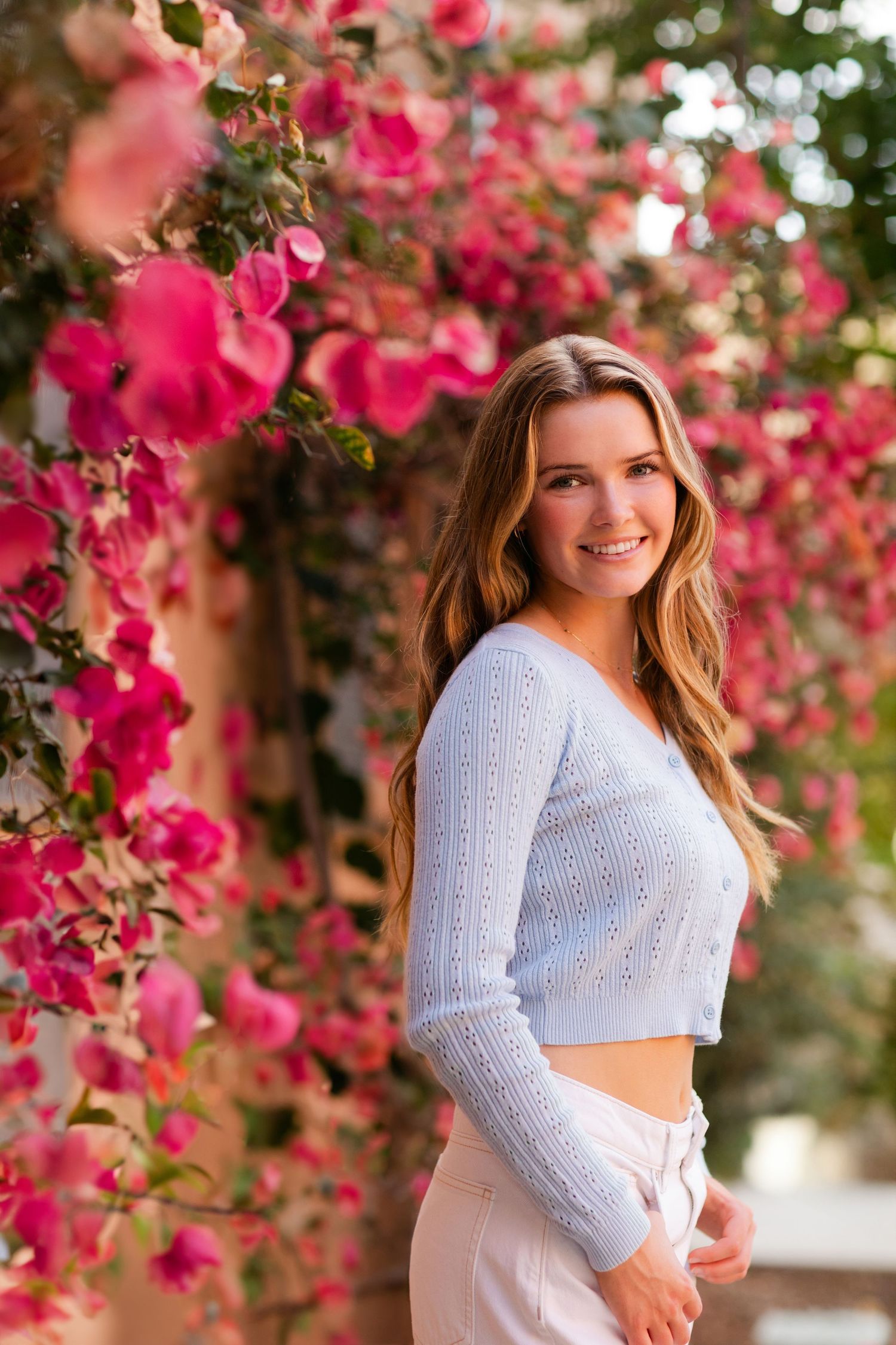 Someone poses in a white cropped sweater against a wall covered in vibrant pink bougainvillea flowers.