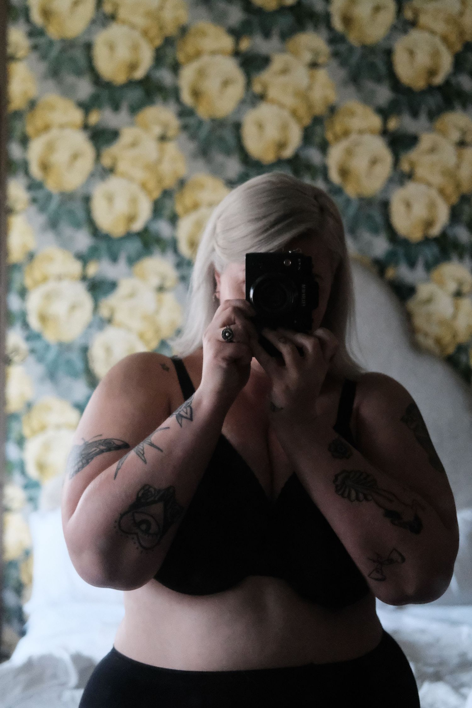 Mirror selfie taken against floral wallpaper showing person in black undergarments with visible arm tattoos.