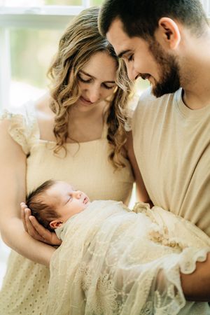 Tender moment with newborn baby wrapped in lacy white blanket as parents gaze lovingly in soft natural light.