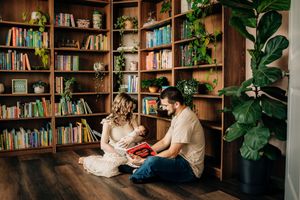 Couple reading together on floor in cozy home library with wooden bookshelves and plants.