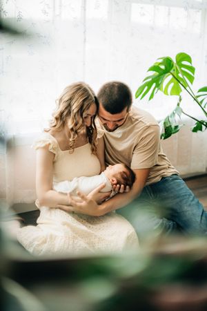 Intimate indoor portrait of parents sharing a gentle moment with their baby near houseplants.