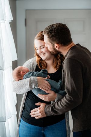 Family moment by window with newborn baby wrapped in grey and blue blankets.