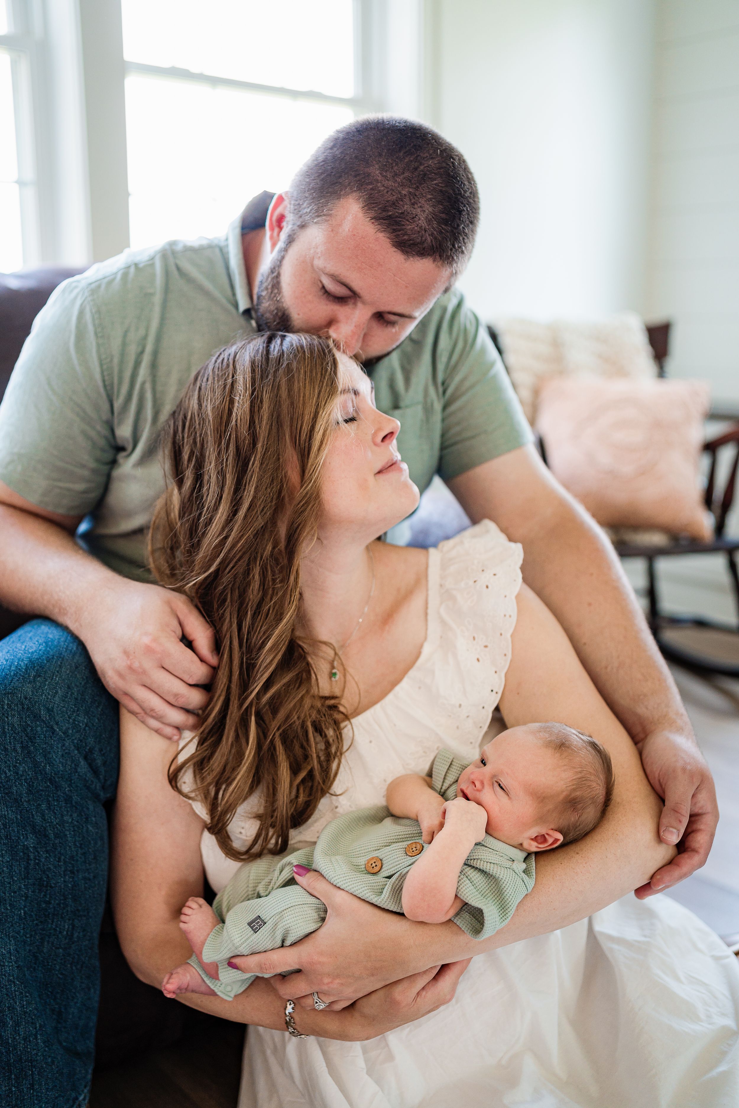Intimate family moment with newborn baby swaddled in mint green being held by parents on a couch.