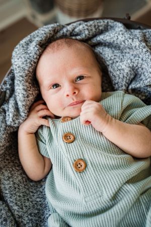 Close up of newborn baby wearing mint green sweater with wooden buttons lying on grey blanket.