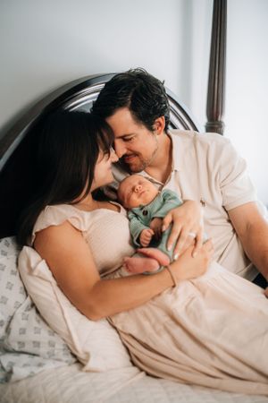 Loving parents cuddling with their newborn baby wearing mint outfit while relaxing in bed.