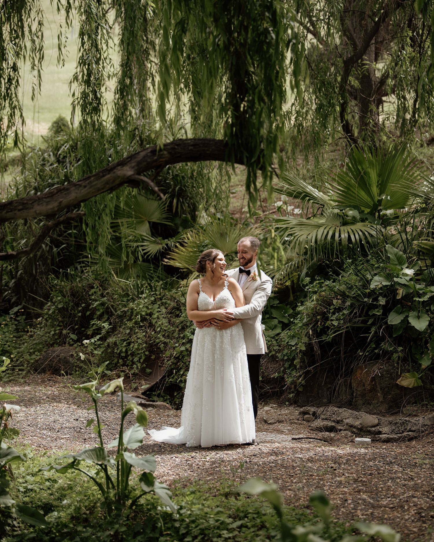 Two people in elegant white dresses share an intimate moment under drooping willow trees in a natural garden setting.