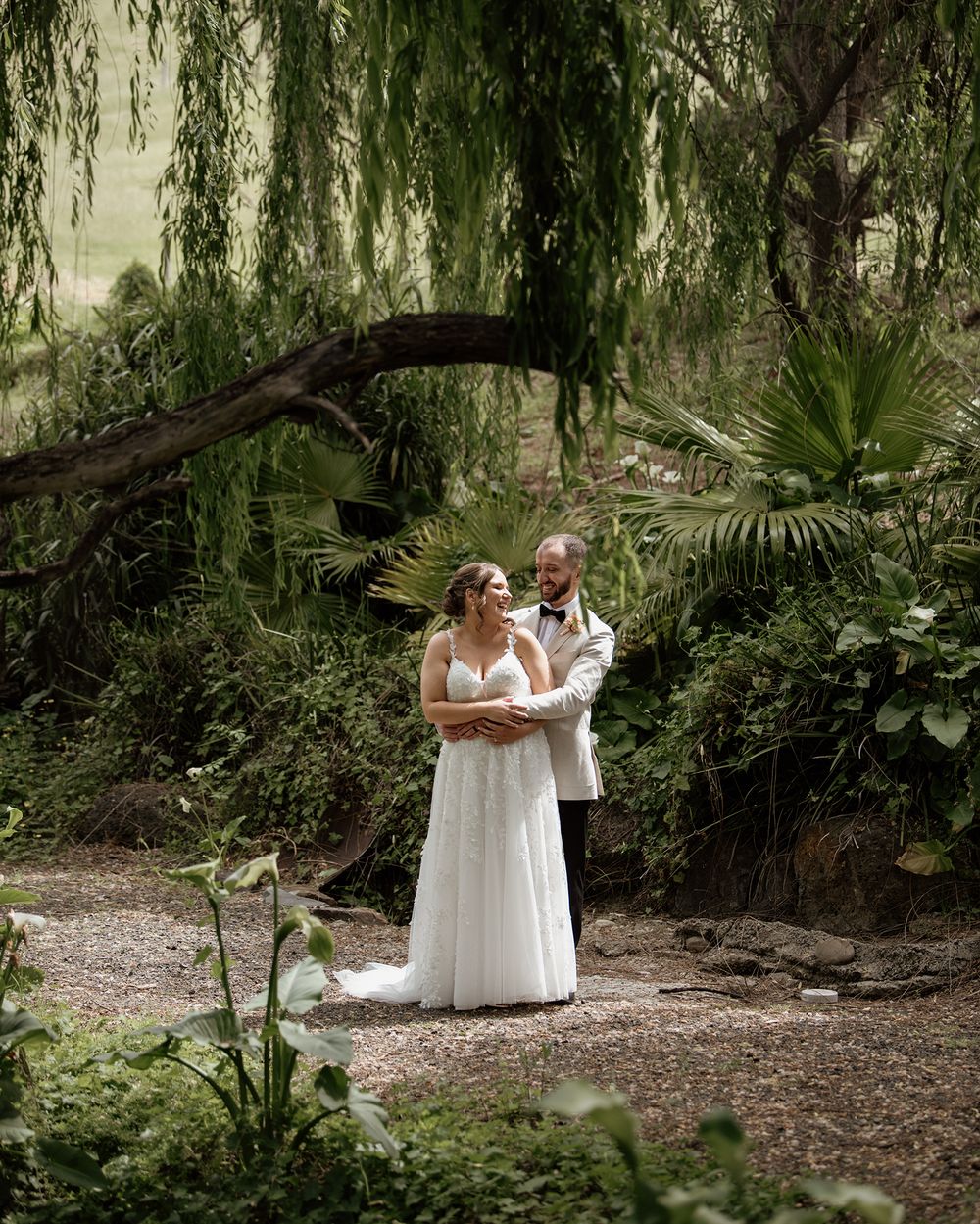 Two people in elegant white dresses share an intimate moment under drooping willow trees in a natural garden setting.