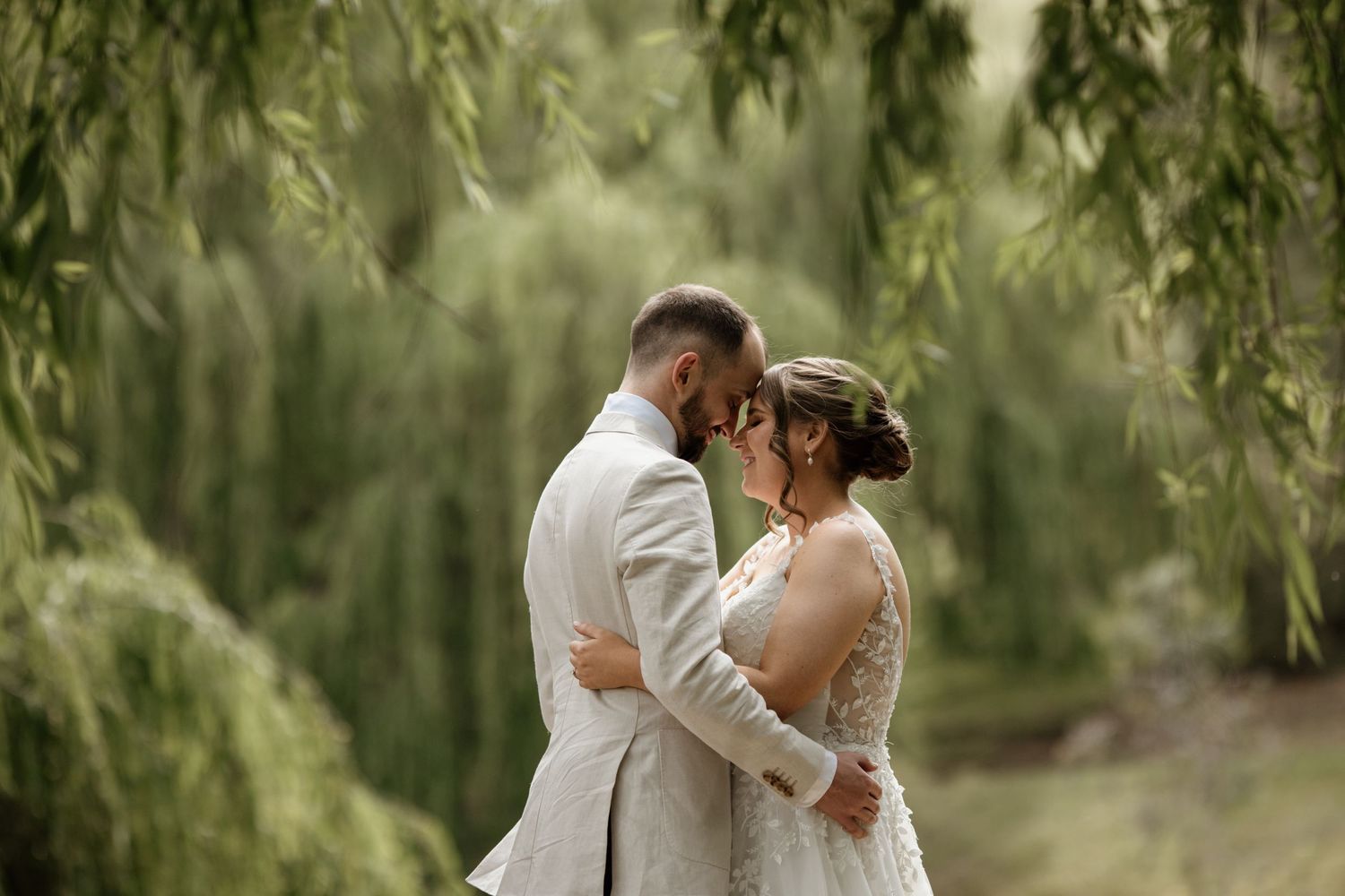 A romantic wedding couple embraces beneath drooping willow branches in a soft, dreamy outdoor setting.