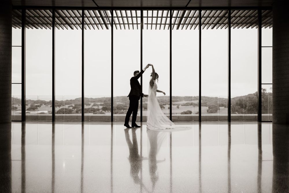 Two silhouettes dance together in an elegant venue with floor-to-ceiling windows overlooking a scenic mountain landscape.
