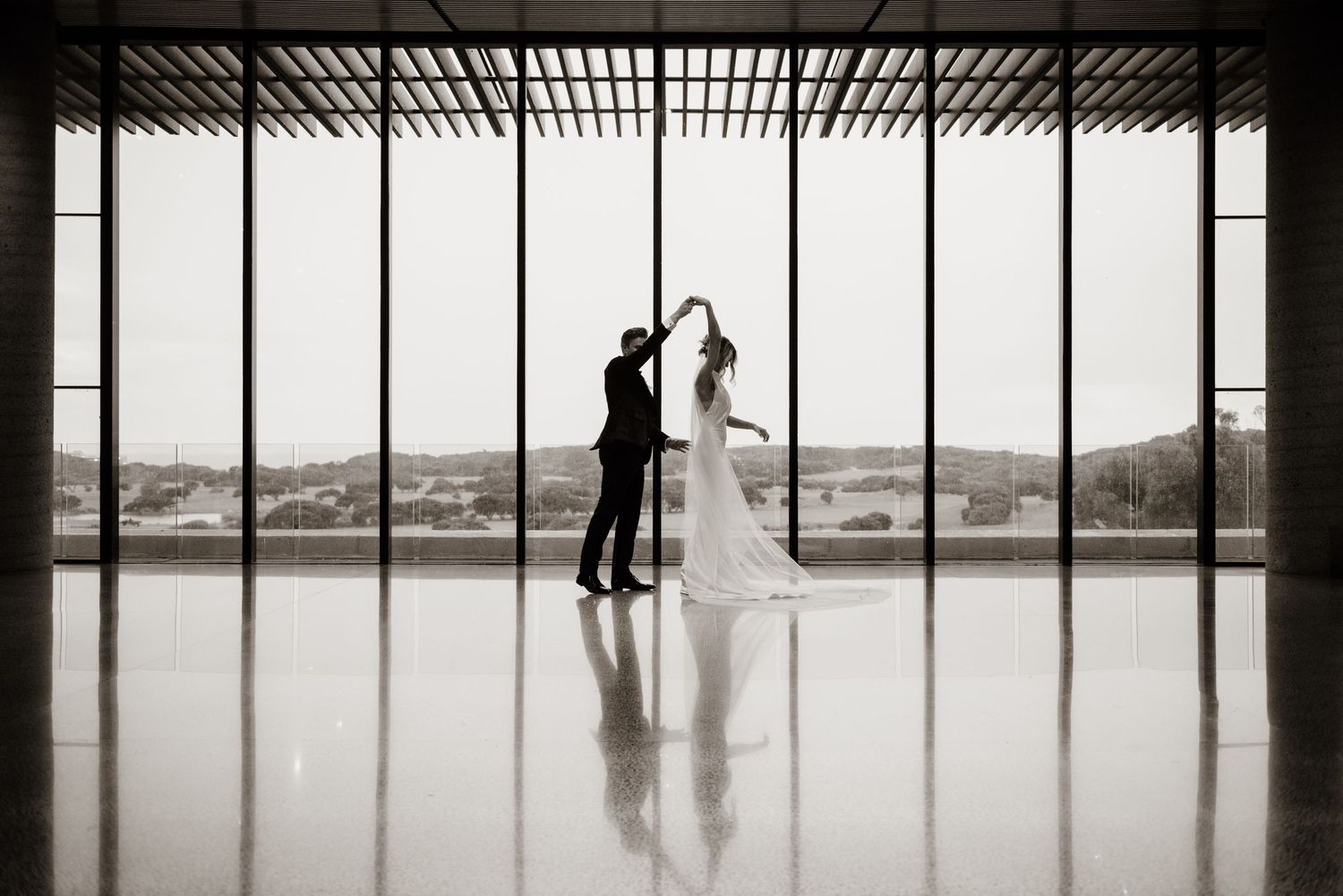 Two silhouettes dance together in an elegant venue with floor-to-ceiling windows overlooking a scenic mountain landscape.
