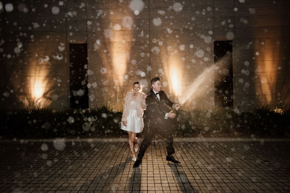 A couple dances joyfully in the snow against illuminated stone pillars at night, creating a magical winter atmosphere.