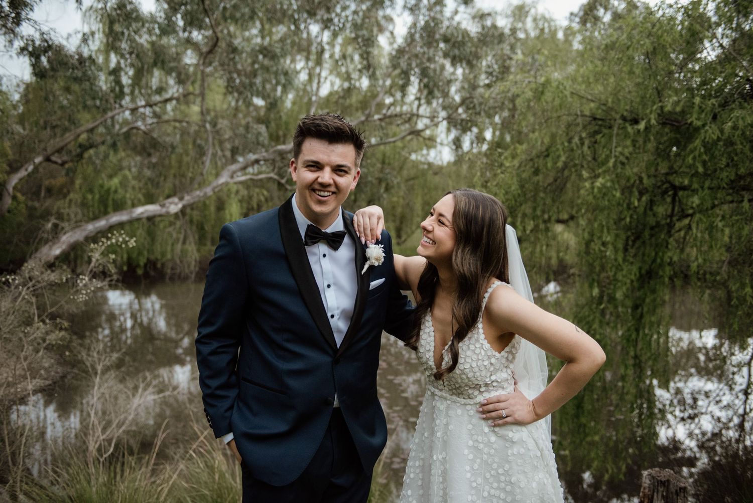 A couple in formal wedding attire embraces and laughs together in front of green trees and natural scenery.